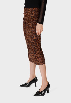 Mujer con falda midi marrón con estampado de leopardo, top negro transparente de manga larga y zapatos de tacón altos negros brillantes con punta puntiaguda, de pie frente a un fondo liso.