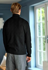 Czarny sweter dzianinowy z teksturowanym wzorem w diamenty, dopasowanym kołnierzem i ściągaczami przy rękawach, w połączeniu z szarymi spodniami w wzory.