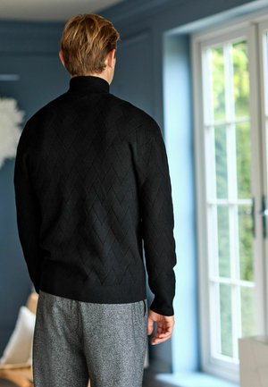 Schwarzer gestrickter Pullover mit einem strukturierten Rautenmuster, enganliegendem Kragen und gerippten Manschetten, kombiniert mit grauen gemusterten Hosen.