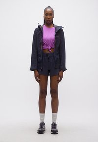 Γυναίκα όρθια, κοιτάζοντας μπροστά, φοράει μωβ τοπ με κόμπο, μαύρο σακάκι και σορτς, λευκές κάλτσες και μαύρα αθλητικά παπούτσια, πάνω σε λευκό φόντο.