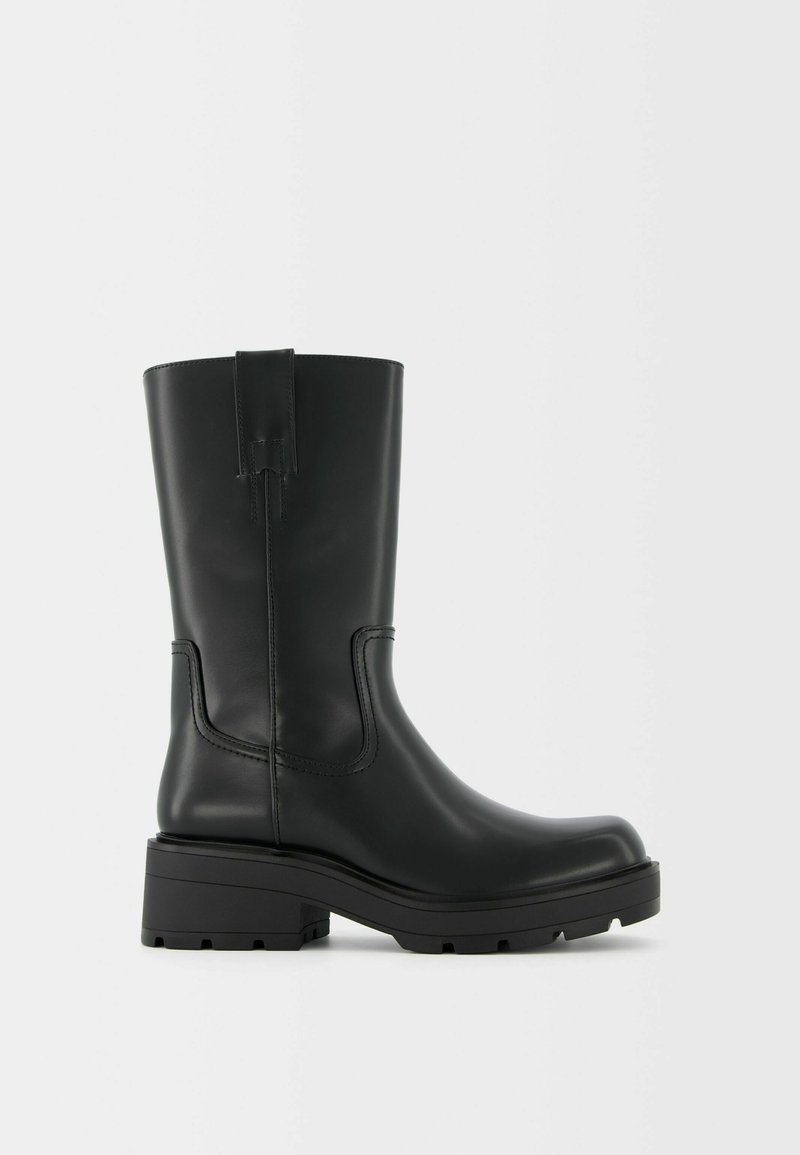 Stivale alla caviglia in pelle nera con suola spessa, punta rotonda e cuciture decorative minime. Presenta un'impugnatura sul retro per facilitare l'indossamento.