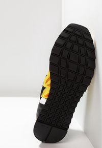 Suela negra con un tread texturizado, presentado un vibrante patrón floral en amarillo y rojo en una parte superior sintética. Acentos de goma visibles.