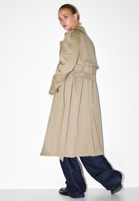 Kobieta w beżowym trenczu, ciemnych szerokich dżinsach i czarnych butach, odwracająca się w trakcie chodzenia na białym tle.