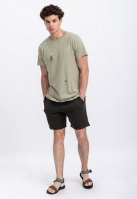 Olijfgroene T-shirt met klimafdruk, gecombineerd met donkere shorts. Model draagt zwart en grijze sandalen. Gladde textuur, casual pasvorm.