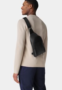 Schwarze Lederumhängetasche mit glatter Oberfläche, ausgestattet mit einer Fronttasche mit Reißverschluss und einem verstellbaren Tragegurt, getragen über einem hellbeigen Pullover.