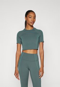 Idrettssett i mørkegrønn stoff, med en kortermet crop-top og turkise sytråder, kombinert med høytliv leggings.