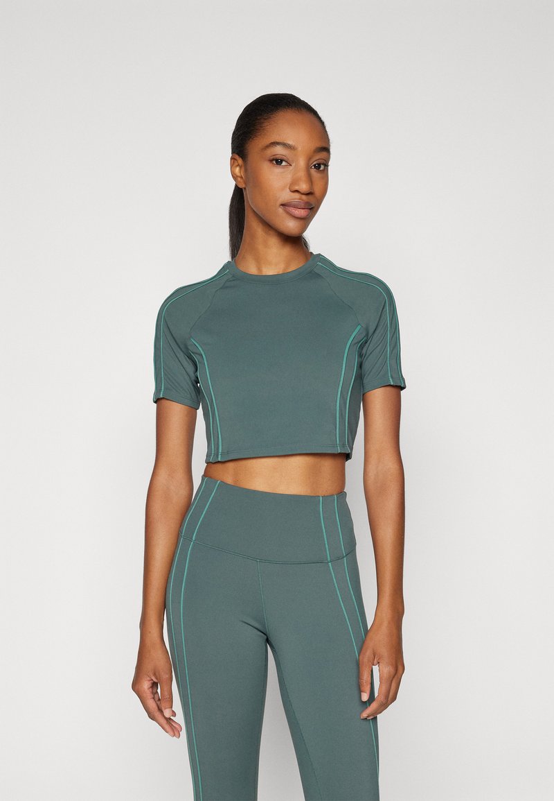 Idrettssett i mørkegrønn stoff, med en kortermet crop-top og turkise sytråder, kombinert med høytliv leggings.
