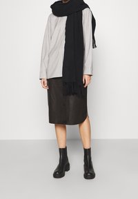 Graues Langarmshirt, schwarzer Wollschal, braune Lederrock und schwarze Ankle Boots mit klobigem Absatz. Minimalistisches Design mit klaren Linien.