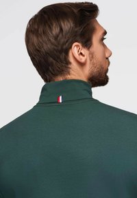 Dunkelgrünes Poloshirt mit hohem Kragen. Verfügt über einen kleinen rot-, weiß- und blauen Akzent am Halsausschnitt. Glatte Textur.