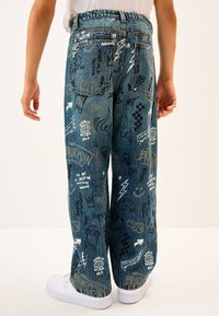 Pantaloni a gamba larga in denim blu con vari disegni grafici bianchi e neri, tra cui testi, fulmini e forme astratte.