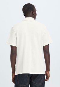 Polo à manches courtes beige clair en tissu texturé, doté d'un col classique et d'un ourlet droit. Design simple et épuré.