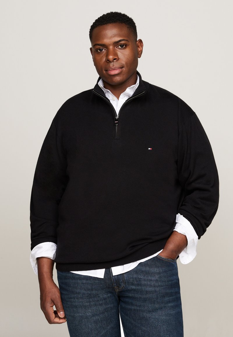 Homme portant un pull noir zippé sur une chemise blanche et un jean bleu, debout avec une main dans la poche devant un fond uni.