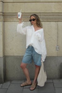 Balta, pārmērīga jēkaba krekls ar uzrullētām piedurknēm, apvienots ar ziliem džinsu šortiem un brūnām sandālēm. Tur rokā dzēriena krūzi, stāvot pret akmens sienu.