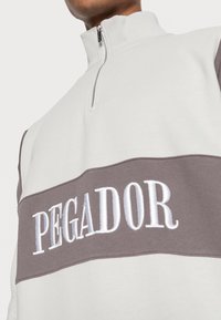 Sudadera blanca con cremallera que presenta una banda horizontal gris con "PEGADOR" bordado en blanco. Tejido suave, cuello alto y cremallera visible.