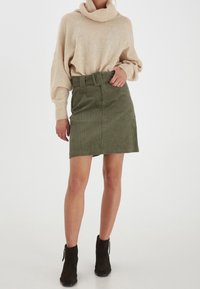 Beige tröja med kjolkrage, i kombination med en grön sammetskjol med bälte. Modellen bär svarta ankelboots med dragkedjedetalj.