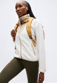 Γυναίκα που φοράει λευκό σακάκι με φερμουάρ, ελιά κολάν και μουσταρδί κίτρινη τσάντα πλάτης, κοιτάζοντας μπροστά με συγκεντρωμένη έκφραση.