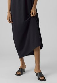 Naine kannab musta midi-kleid ja musti avatud varbaga kontsakingi, tõstab kleiti ühe käega veidi üles.
