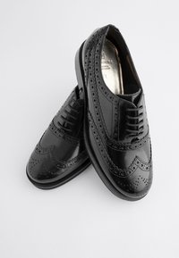 Scarpe in pelle nera con design brogue, punta arrotondata e dettagli in rilievo sui lati. Chiusura con lacci e finitura liscia.