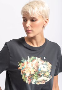 Grijze katoenen t-shirt met een kleurrijk grafisch ontwerp waarop mensen, meubels en planten zijn afgebeeld, in een surrealistische, gelaagde compositie.