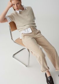 Maglione di lana beige indossato sopra una camicia bianca, abbinato a pantaloni di velluto a coste color sabbia e mocassini marrone scuro, seduto su una sedia in metallo cromo.