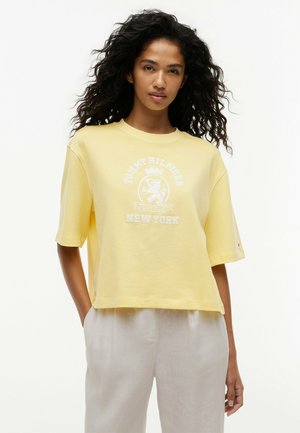 Kobieta z długimi kręconymi włosami nosi żółtą koszulkę Tommy Hilfiger i jasne spodnie, stoi z jedną ręką w kieszeni na białym tle.