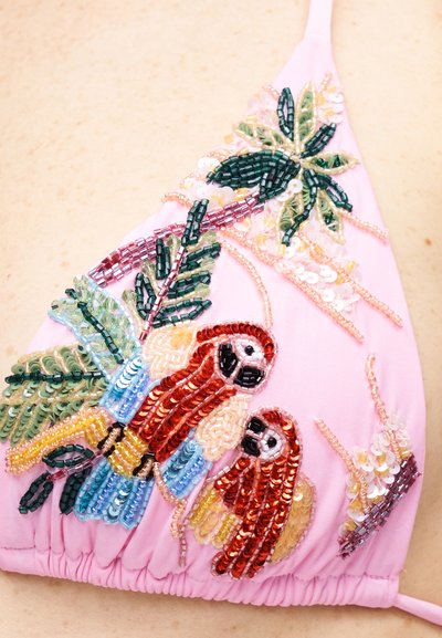 Tissu rose avec une broderie détaillée de perles et de sequins représentant deux perroquets colorés perchés sur des feuilles et des branches vertes.