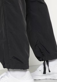 Underben klädda i svarta byxor med justerbara snörningar vid fållarna, kombinerat med vita träningsskor och ribbade vita strumpor.