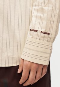 Μπεζ πουκάμισο με κάθετες καφέ ρίγες, με κεντημένο το "SAMSØE" στη μανσέτα. Το ύφασμα φαίνεται ελαφρύ και λείο.