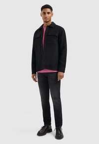 Zwarte wollen shirtjack met twee borstzakken, gedragen over een roze T-shirt, gecombineerd met een donkere spijkerbroek en zwarte veterschoenen.