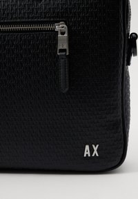Sac noir texturé avec un motif de logo gaufré, quincaillerie de fermeture éclair en argent, et un accent de logo sur le côté, présentant une finition lisse et une forme robuste.