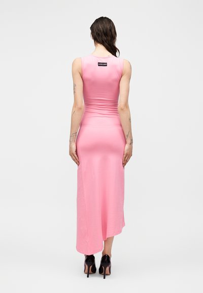 Γυναίκα με τατουάζ που φοράει ροζ εφαρμοστό μίντι φόρεμα χωρίς μανίκια και μαύρα ψηλοτάκουνα, όρθια με την πλάτη προς τα εμπρός.