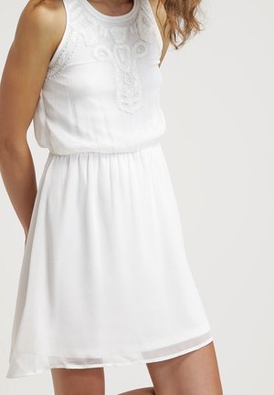 Ärmelloses weißes Kleid mit bestickten Spitzenapplikationen am Oberteil und einem gerafften Taillenbund, getragen von einer stehenden Person vor einfarbigem Hintergrund.