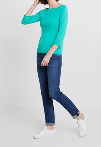 Türkisfarbener, figurnaher Langarmoberteil mit Rundhalsausschnitt, kombiniert mit dunkelblauen Skinny-Jeans und weißen Sneakers. Schlichte und glatte Textur.
