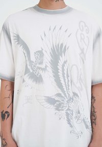 Přehnaně velké bílé tričko s šedými grafickými motivy ptáků, s kulatým výstřihem a krátkými rukávy; hladká textilie.