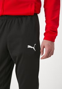 Svart treningsbukser med en hvit Puma-logo på venstre lår. Materialet er glatt med elastisk midjebånd og sidelommer. Rød jakke delvis synlig.