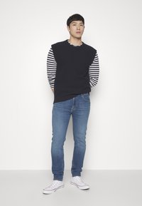 Navy sleeveless sweater over a striped long-sleeve shirt, paired with blue jeans and white sneakers; smooth textures, casual fit.