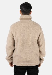 Chaqueta de forro polar beige con cuello alto, presenta una tela texturizada y un ajuste relajado, diseñada para ofrecer calidez y comodidad.