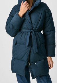 Mörkblå pufferjacka med bälte i midjan, stor huva och dragkedja framtill. Har kviltad textur och sidofickor.