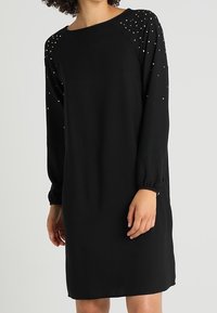 Robe noire à manches longues avec une coupe ample, dotée d'un col rond et d'épaules embellies de petits accents argentés. Matière douce.