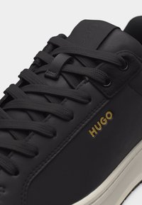 Baskets en cuir noir avec lacets noirs, logo "HUGO" doré sur le côté et semelle en caoutchouc blanche, montrant la partie supérieure en détail.