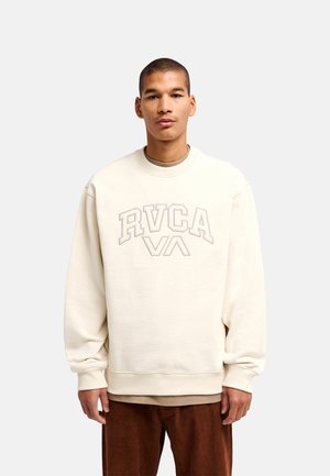 Homme portant un sweat-shirt RVCA couleur crème et un pantalon marron, debout devant un fond clair uni.