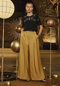 Top negro con embellishments de estrellas doradas, pantalones de pierna ancha dorados de cintura alta. Los accesorios incluyen pendientes de aro y una pulsera.