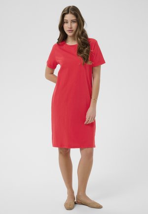 Frau mit langem, welligem Haar, die ein rotes, knielanges Kurzarmkleid und beige geflochtene flache Schuhe trägt, vor einem schlichten Hintergrund steht.