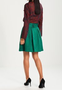 Gonna plissè verde con una finitura testurizzata, abbinata a una blusa rossa a fantasia e tacchi alti neri. La vista posteriore evidenzia la struttura della gonna.