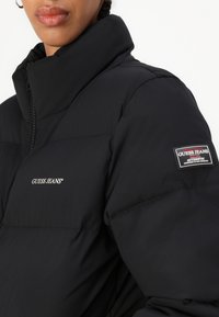Juodas pūkinis pilotas su aukštu apykakle, tekstūruotu siuvinėjimu ir Guess Jeans logotipu ant krūtinės ir rankovės. Lygus audinio apdaila.