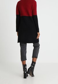 Osoba ubrana w długi czerwono-czarny sweter, z podwiniętymi ciemnoszarymi spodniami i czarnymi, wysokimi botkami na obcasie, stojąca na tle jednolitej ściany.