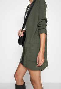 Olivově zelený svetr na knoflíky s texturovaným pletením, otevřený vpředu, dlouhými rukávy, dvěma předními kapsami a volným střihem, nošený přes tmavé šaty.