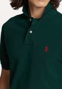 Ciemnozielona koszulka polo wykonana z teksturowanej bawełny, z krótkim kołnierzykiem, dwoma guzikami oraz haftowanym czerwonym logo na piersi.