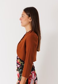 Giacca corta marrone con maniche a tre quarti indossata sopra un vestito a stampa floreale multicolore, che mette in mostra un tessuto morbido e una vestibilità slim.