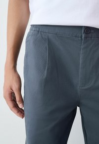 Szürke, könnyű anyagú nadrág sima textúrával, rugalmas derékpánttal és egy gombos zárással; oldalsó zsebekkel és testhezálló fazonnal rendelkezik.